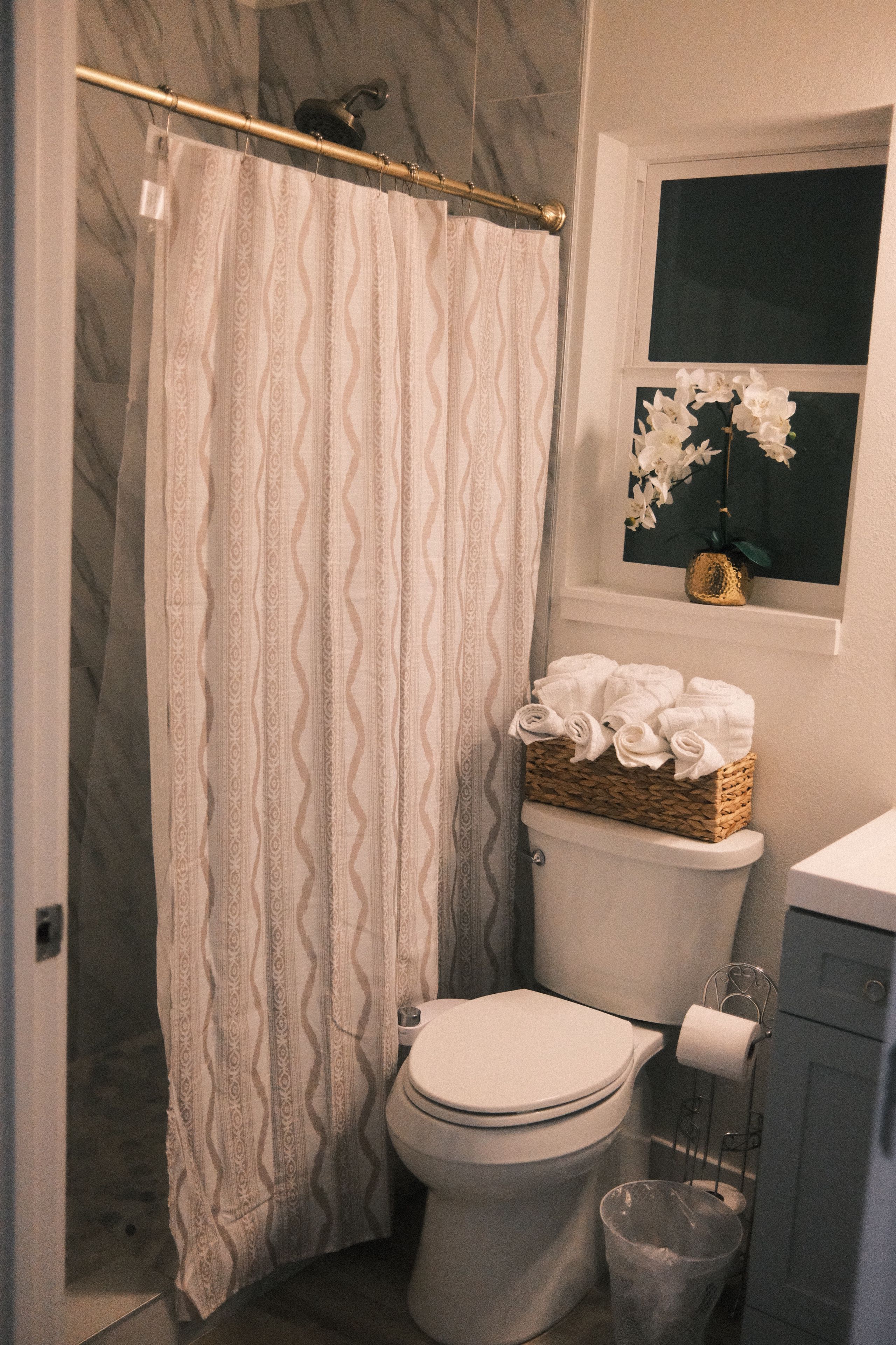 Een badkamer met douchegordijn, toilet, een mand met handdoeken en een raam met een orchidee.