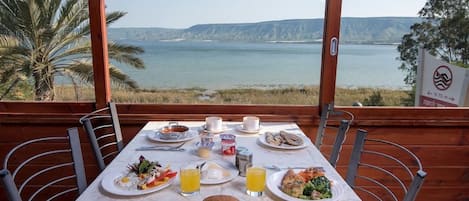 A dining table set with plates of food, glasses of juice, and a view of a lake and hills.