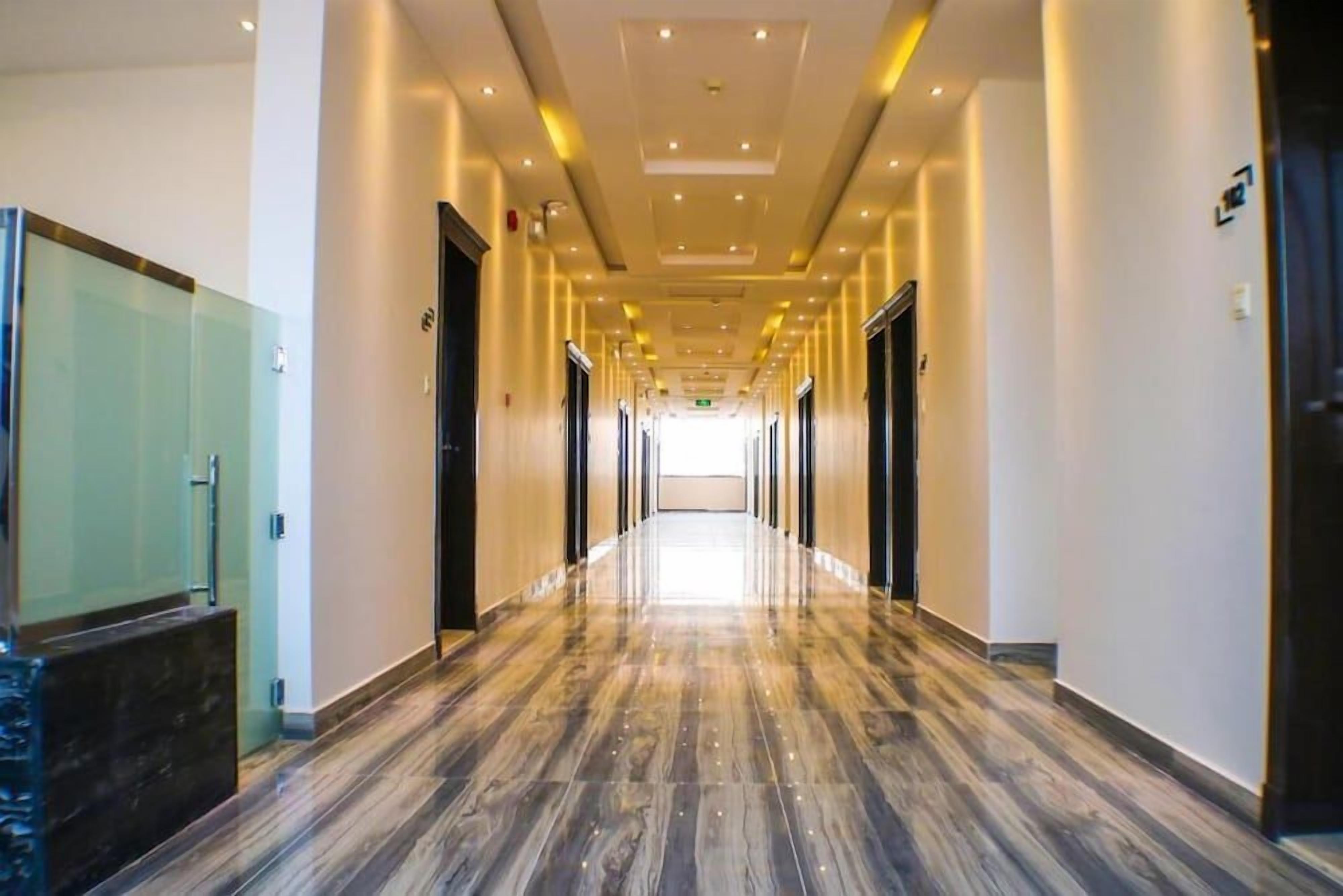 A hotel corridor with a long, polished wooden floor, glass doors on the left, and a bright ceiling with recessed lighting.