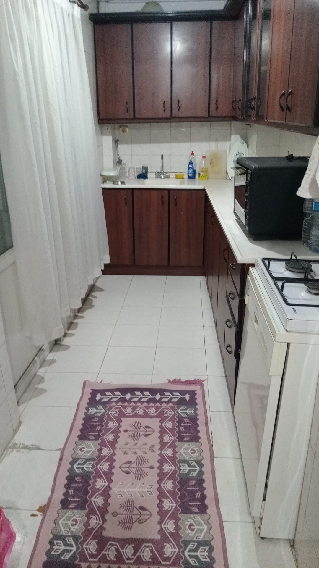 Cocina compacta con armarios de madera, encimera blanca, estufa a gas y una alfombra estampada.