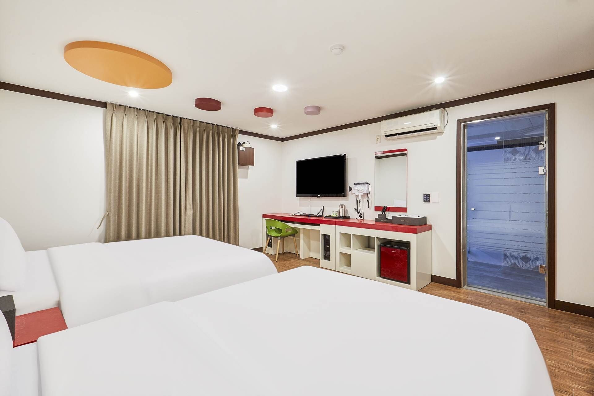 Habitación de hotel con dos camas, un escritorio, un televisor de pantalla plana, un espejo, un equipo de aire acondicionado y una puerta con panel de vidrio esmerilado.