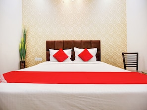 Habitación de hotel con cama, dos almohadas rojas, cabecero de madera, una silla y una planta en maceta.