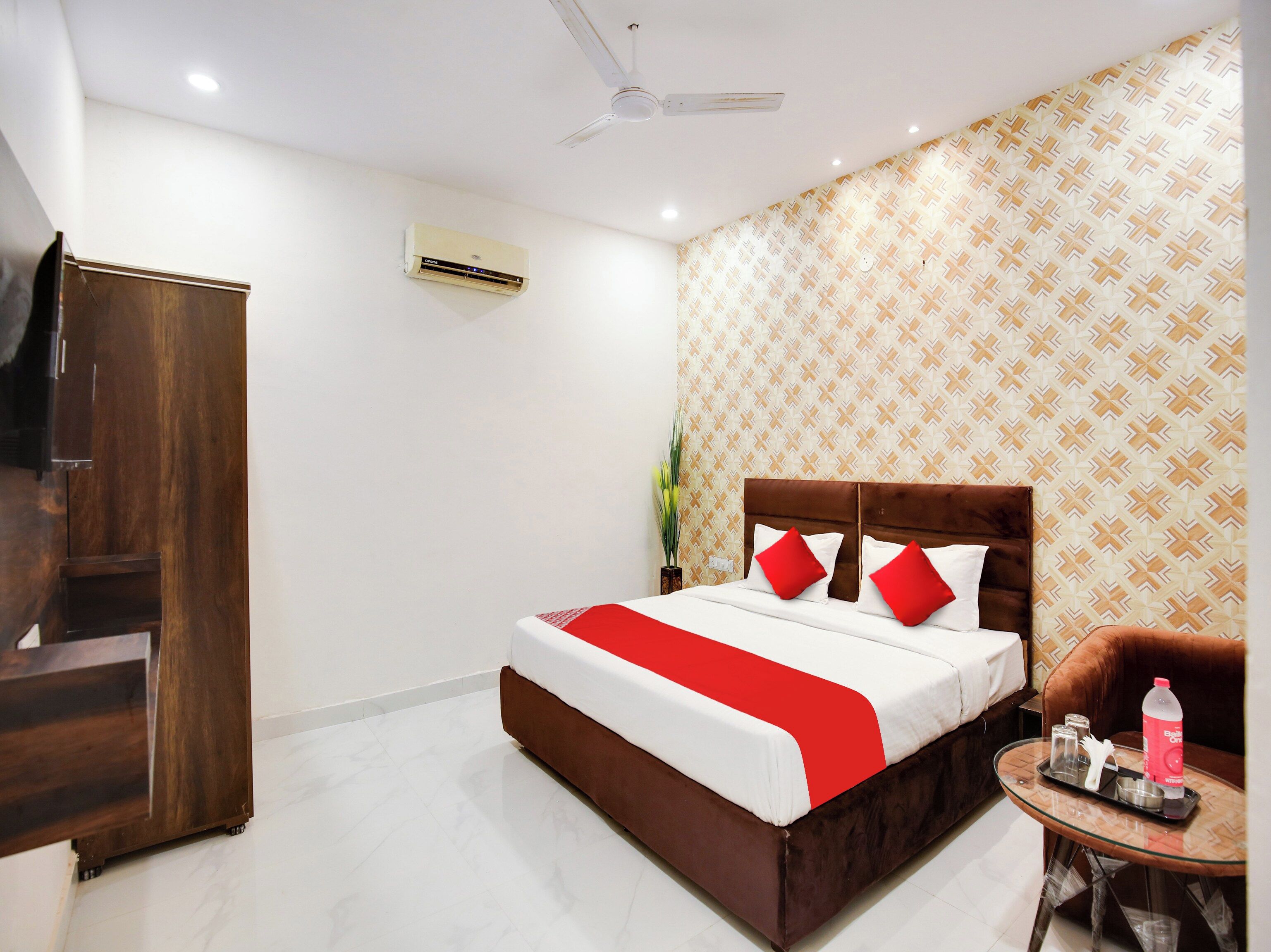 A hotel room with a bed, a TV, a ceiling fan, a patterned wall, and a small table with a bottle and glasses.