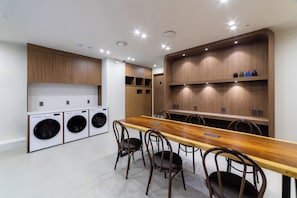 A modern laundry room with a long wooden table and chairs, a washing machine, and a shelf with bottles.