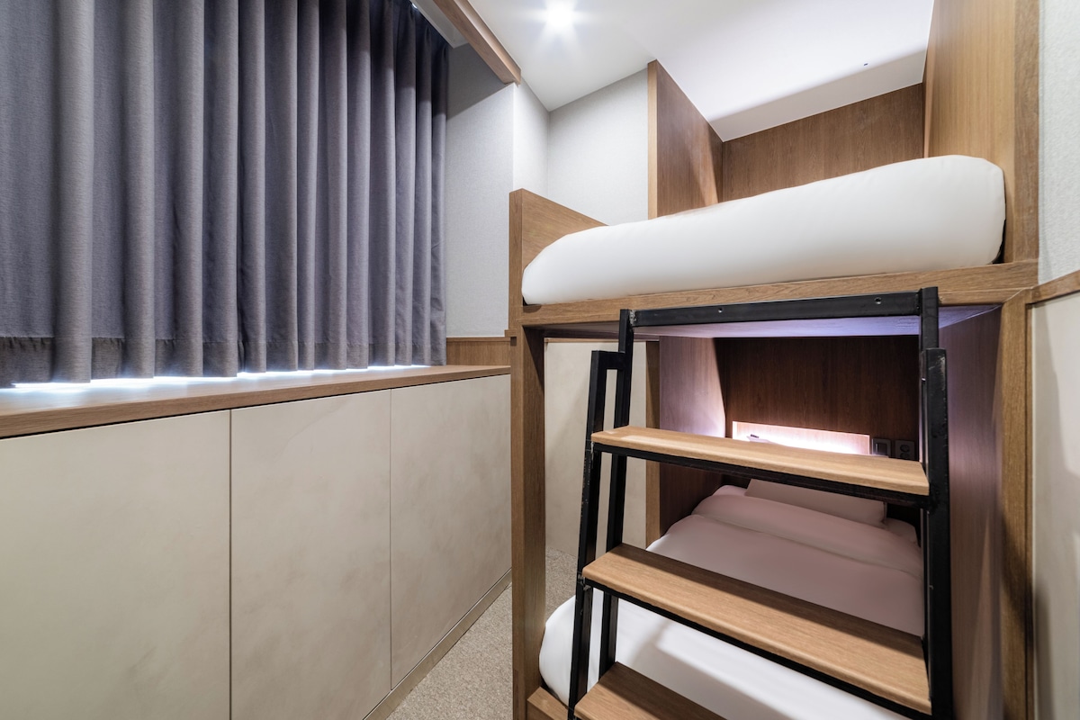 A bunk bed with a wooden frame and white bedding, a window with grey curtains, and a light fixture on the ceiling.