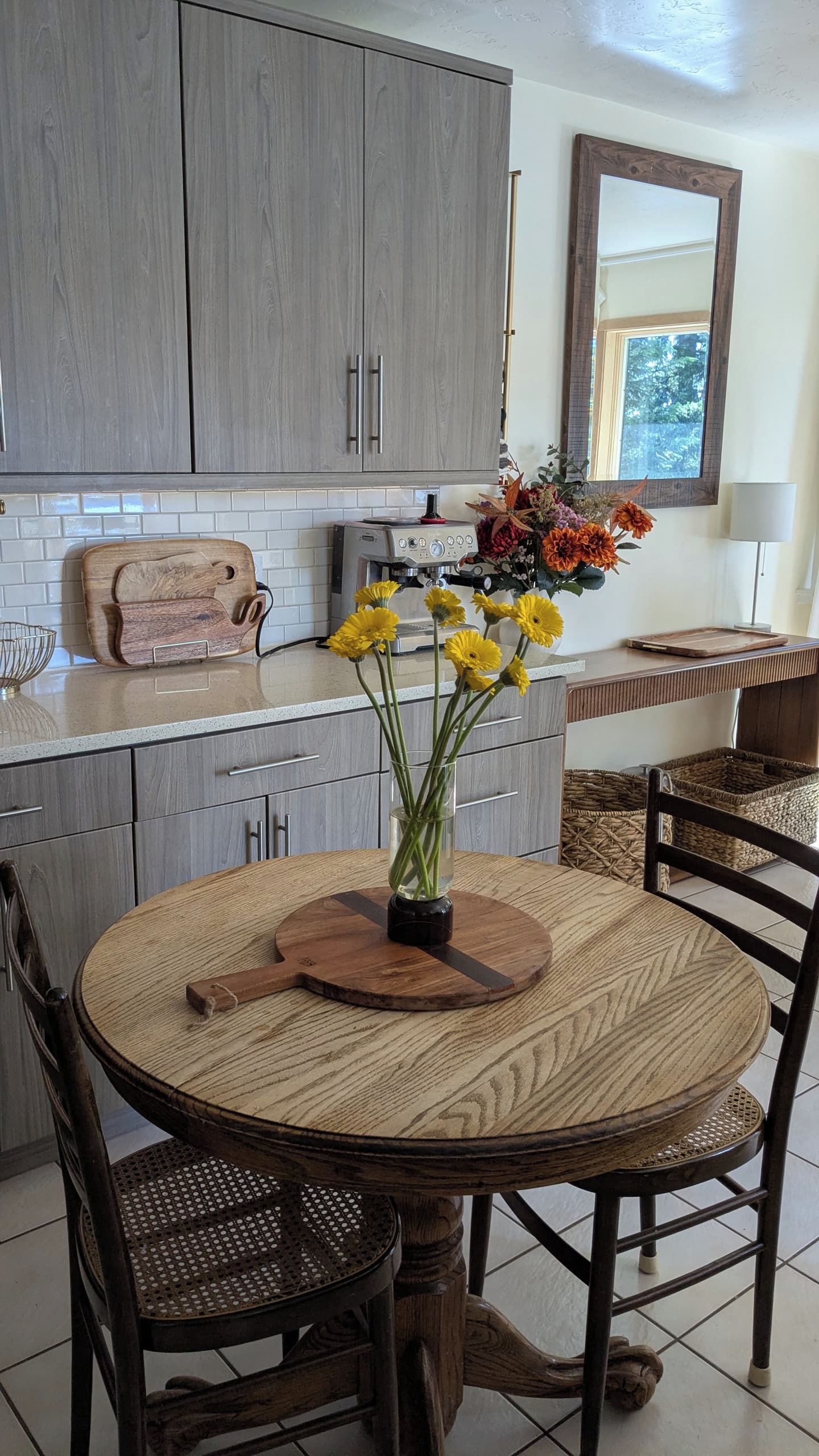 A kitchen with grey cabinets, a coffee machine, a vase with yellow flowers, and a round wooden table with chairs.