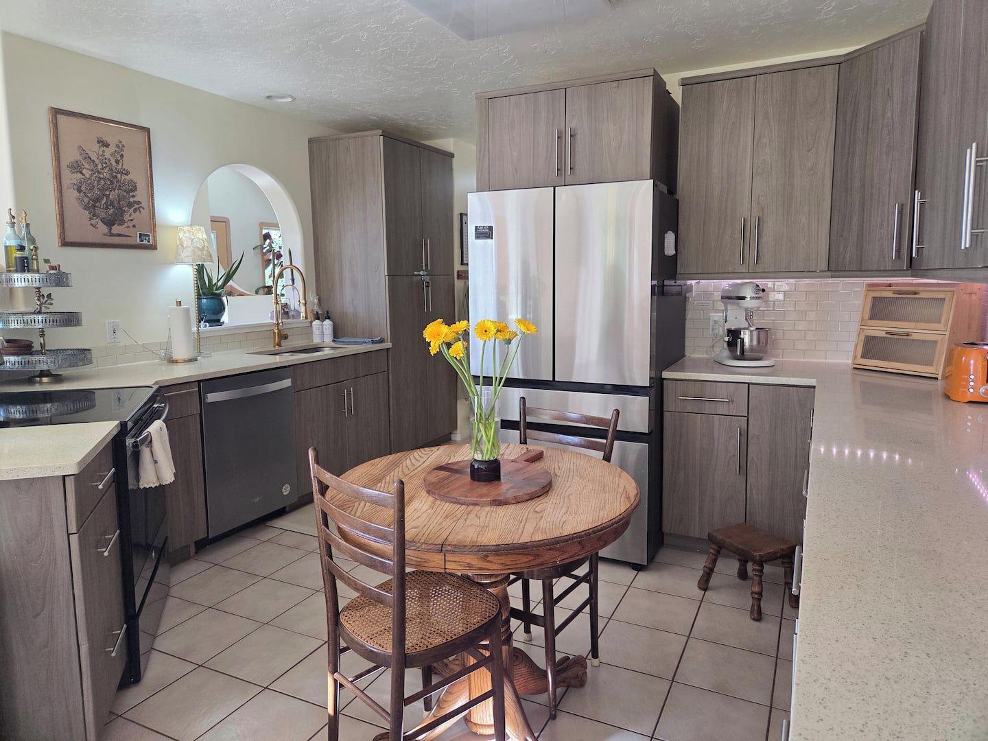 A modern kitchen with wooden cabinets, a stainless steel refrigerator, a dining table with chairs, and a vase with flowers.