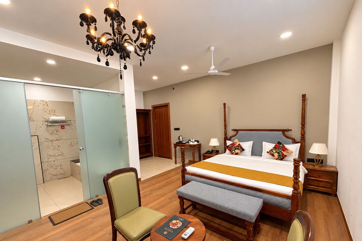 Ein Hotelzimmer mit Bett, Schreibtisch, Stuhl, Bank, einer Kristallleuchter und einem Deckenventilator, sowie einem Badezimmer mit Dusche und Toilette.