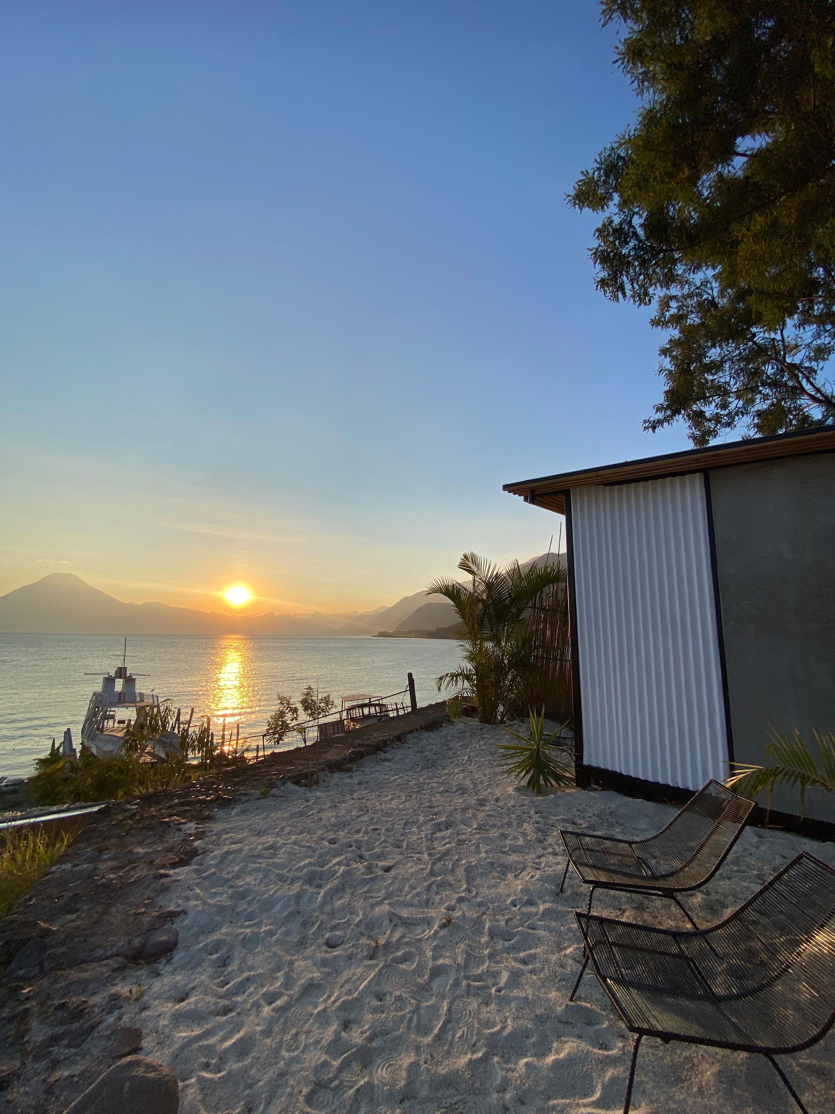 Un patio frente a la playa con vistas al mar y a las montañas, que incluye un bote pequeño, una zona de estar cubierta y una estructura de madera.