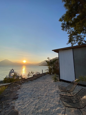 Un patio frente a la playa con vistas al mar y a las montañas, que incluye un bote pequeño, una zona de estar cubierta y una estructura de madera.