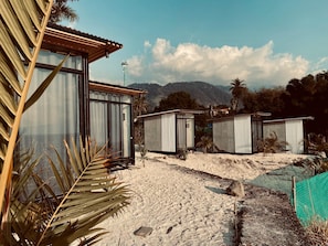 Una propiedad frente a la playa con varias cabañas de playa de estructura metálica, un edificio de madera con techo inclinado y montañas al fondo.