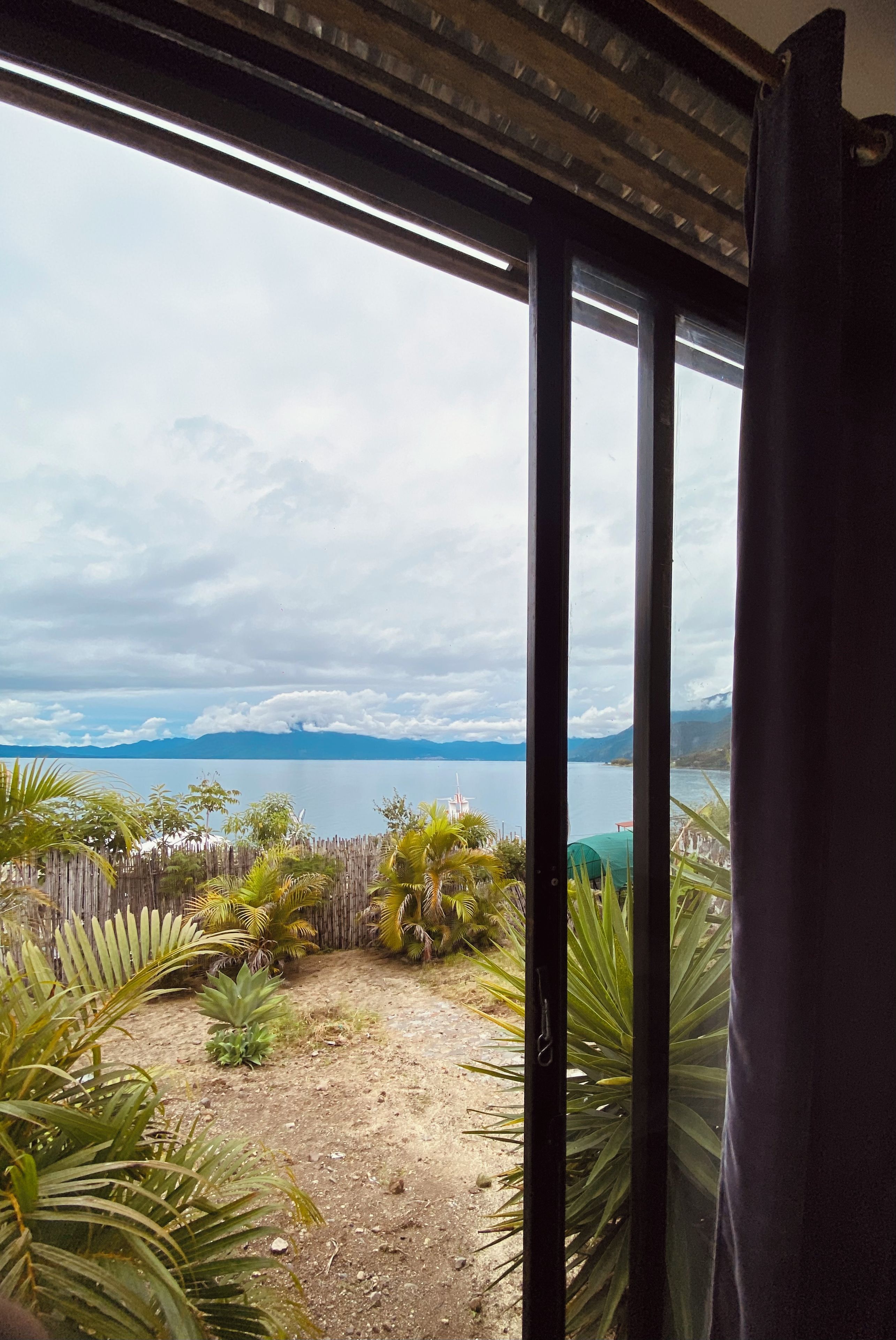 Vista a través de una ventana con marco metálico, que muestra un paisaje natural con una masa de agua, montañas al fondo y diversa vegetación, incluyendo palmeras.