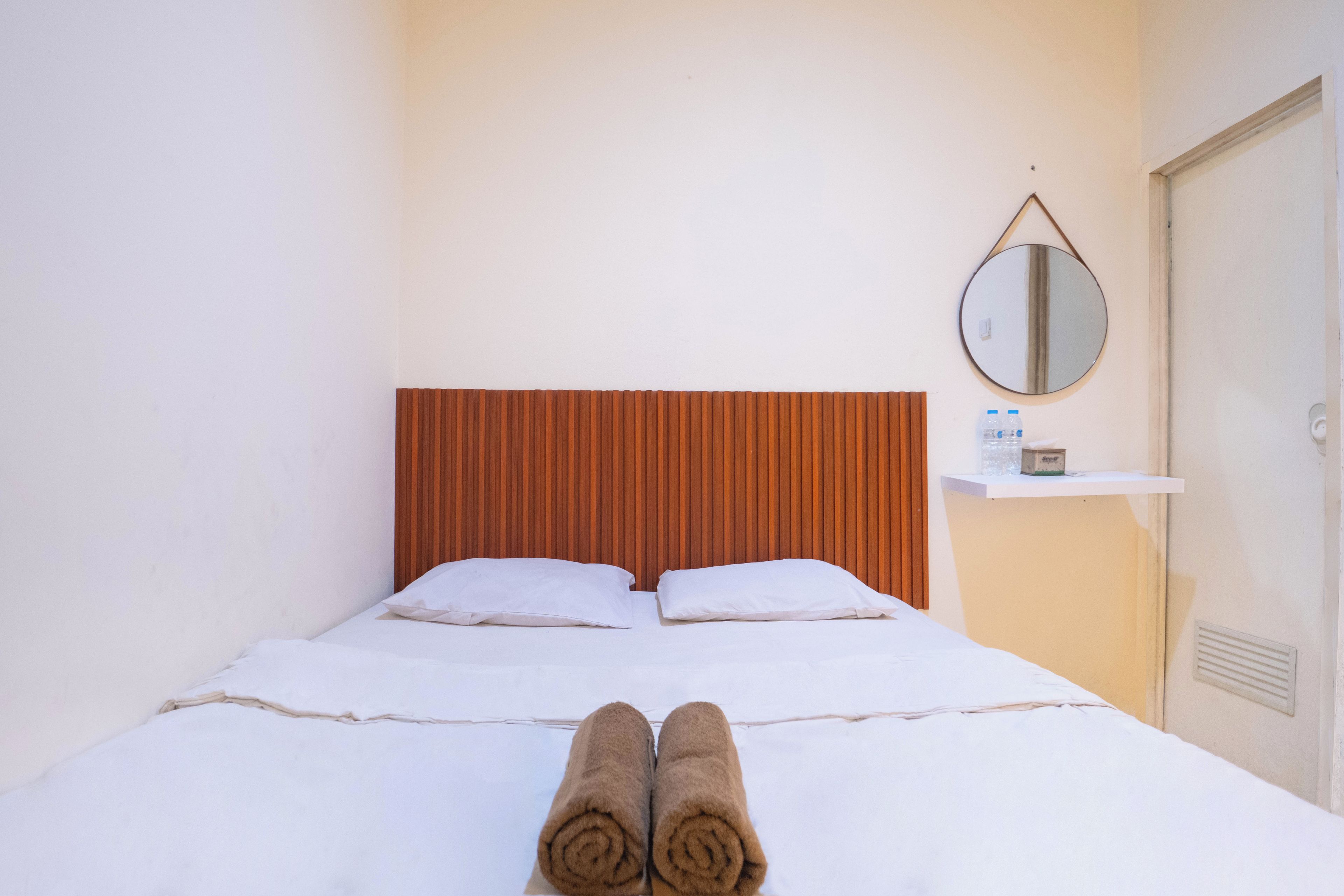 A hotel room with a bed, pillows, a wooden headboard, a bedside table with a bottle and a box, and a round mirror on the wall.