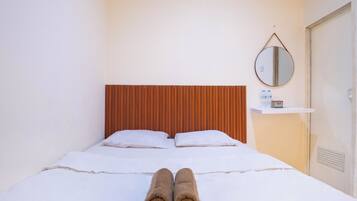 A hotel room with a bed, pillows, a wooden headboard, a bedside table with a bottle and a box, and a round mirror on the wall.