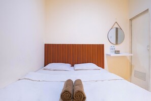 A hotel room with a bed, pillows, a wooden headboard, a bedside table with a bottle and a box, and a round mirror on the wall. - Tamastu House Grogol (Jakarta Barat)