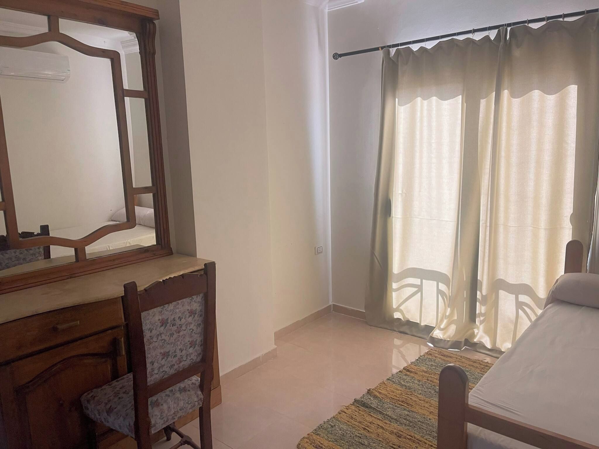 A bedroom with a bed, a wooden desk with a chair, a large mirror, and a window with curtains.