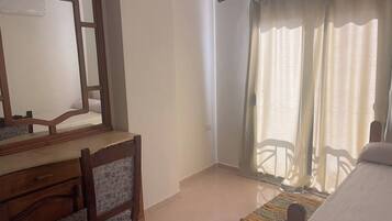 A bedroom with a bed, a wooden desk with a chair, a large mirror, and a window with curtains.
