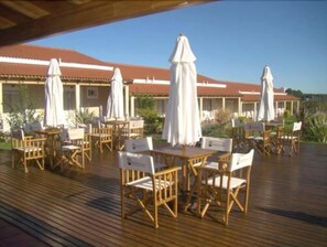 Een buitenruimte met houten tafels en stoelen, witte parasols en een gebouw met een rood dak op de achtergrond.