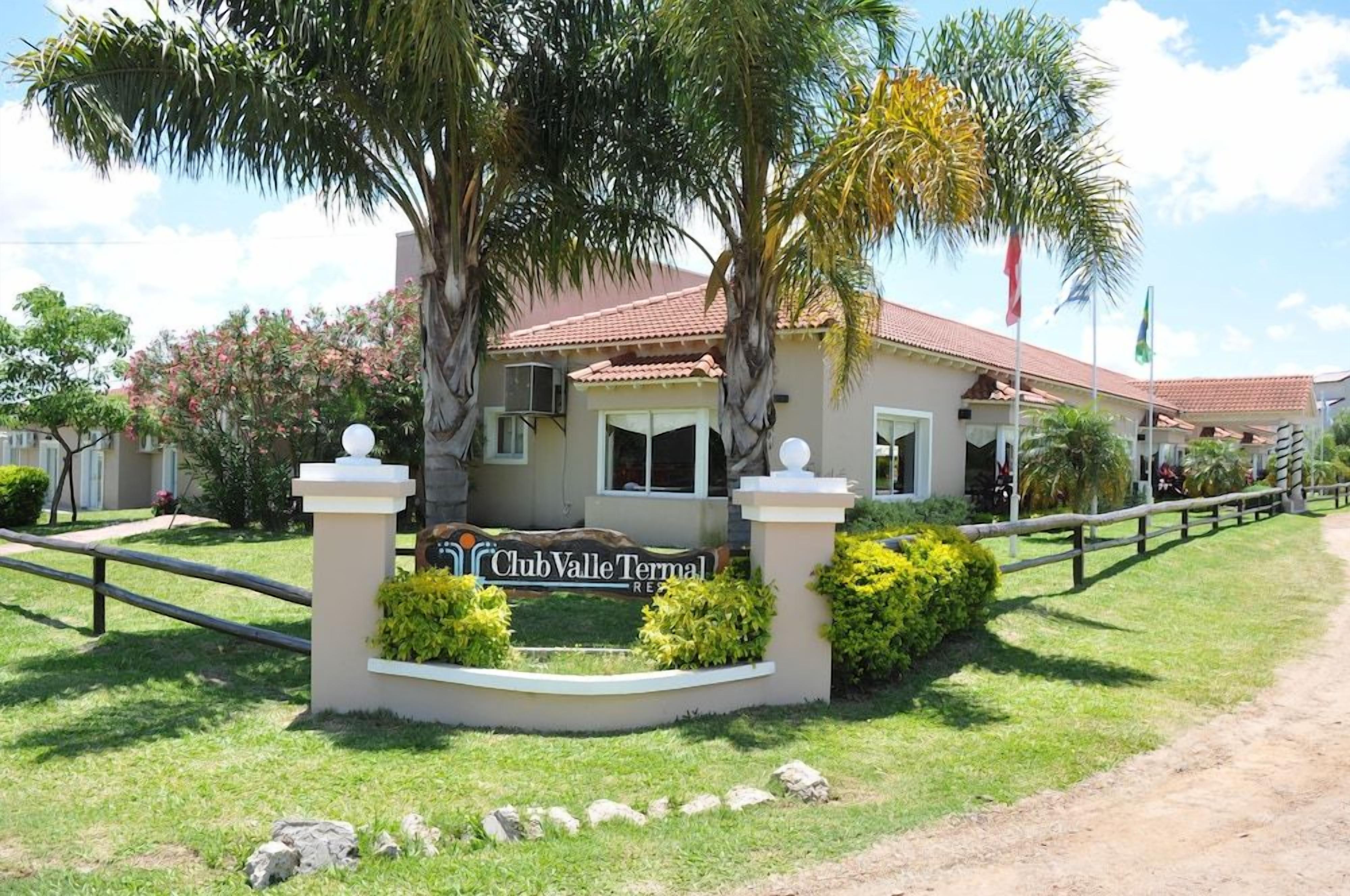 Complejo turístico Club Valle Termas con un cartel, palmeras y un sendero cercado.