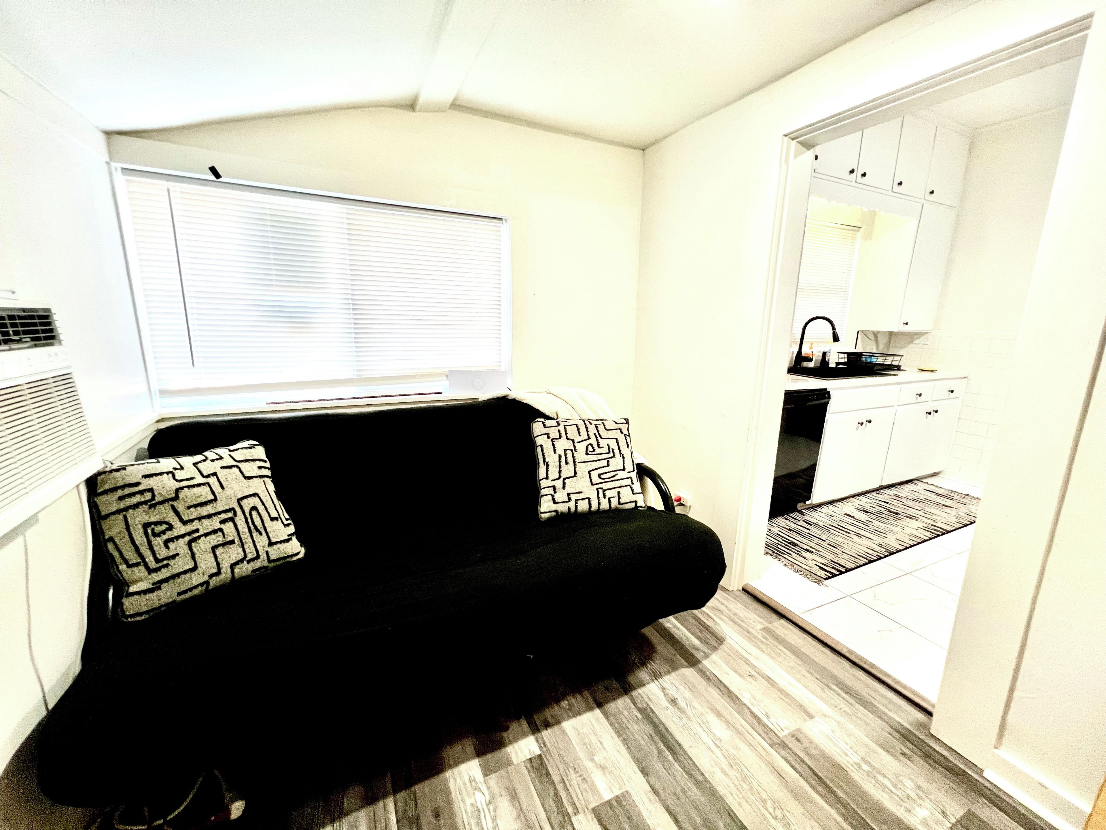 A compact living space with a sofa, patterned pillows, a window with blinds, and a kitchenette area.
