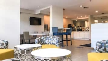 A modern hotel lobby with round tables, comfortable seating, a flat-screen TV, and a visible kitchen area.
