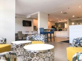 A modern hotel lobby with round tables, comfortable seating, a flat-screen TV, and a visible kitchen area.