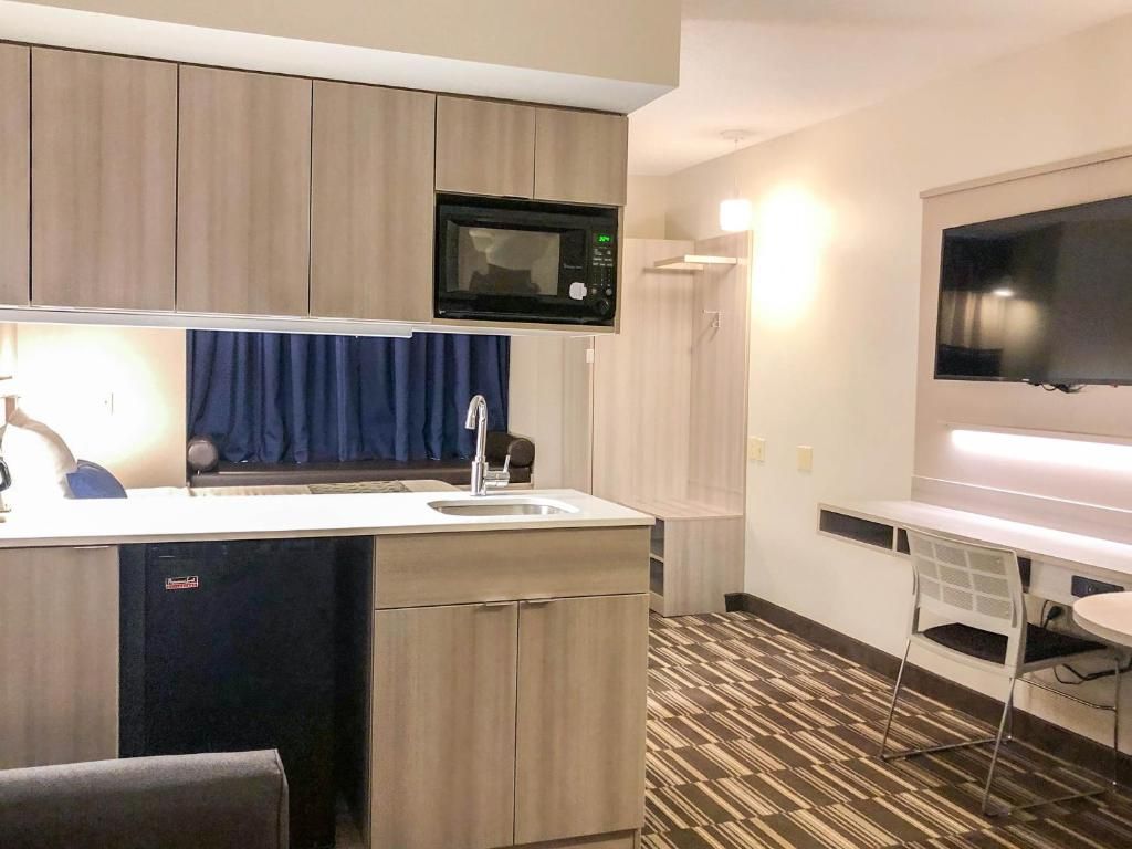 Habitación de hotel compacta con kitchenette, televisor de pantalla plana, una pequeña mesa de comedor y alfombra estampada.