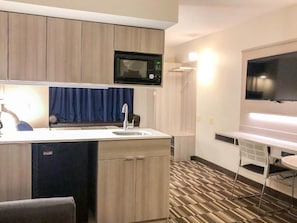 Habitación de hotel compacta con kitchenette, televisor de pantalla plana, una pequeña mesa de comedor y alfombra estampada.