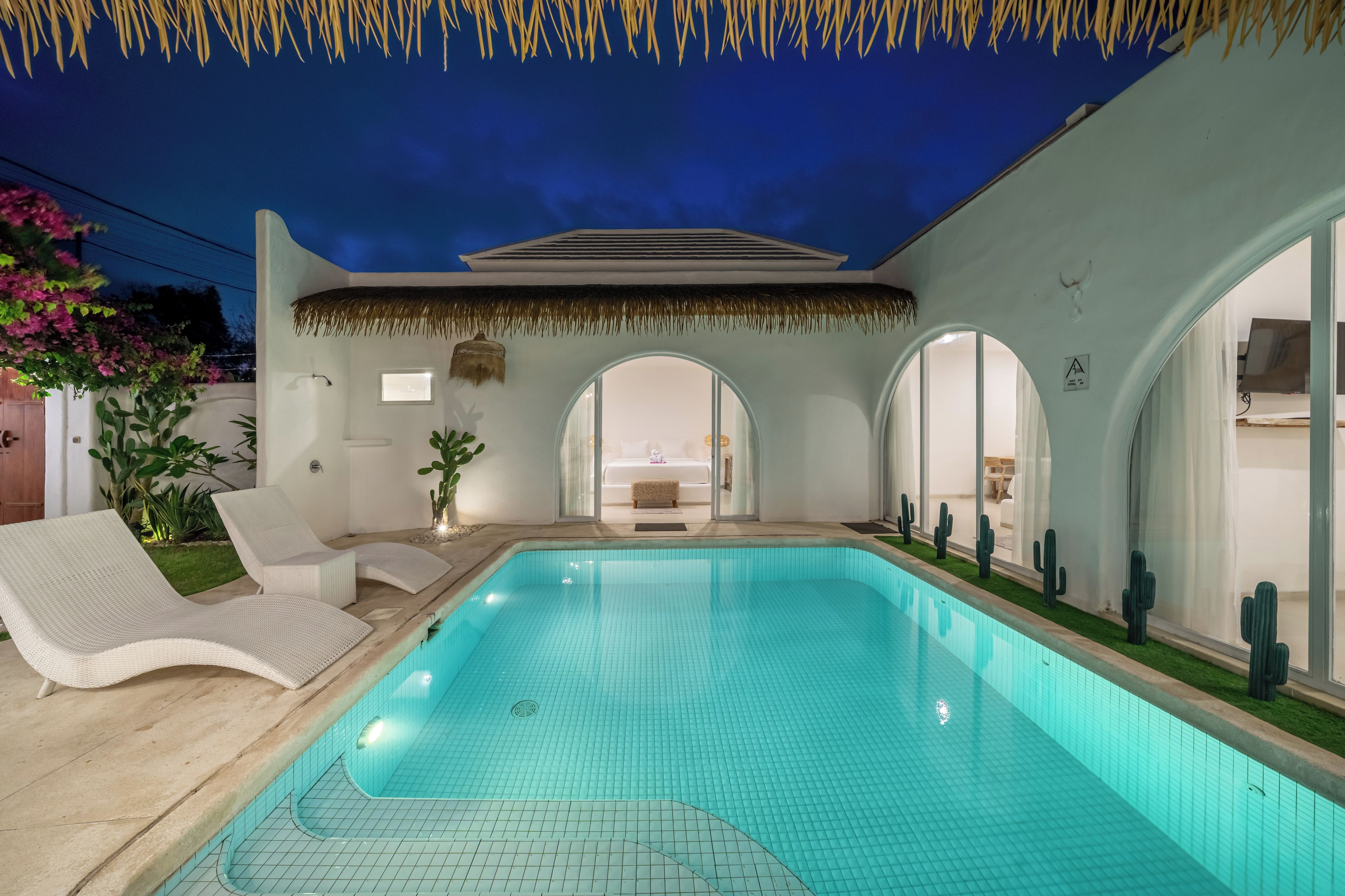 Una piscina con solárium de baldosas, una silla de descanso y una estructura de techo de paja.