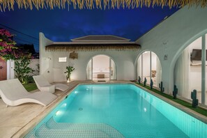 Una piscina con solárium de baldosas, una silla de descanso y una estructura de techo de paja.