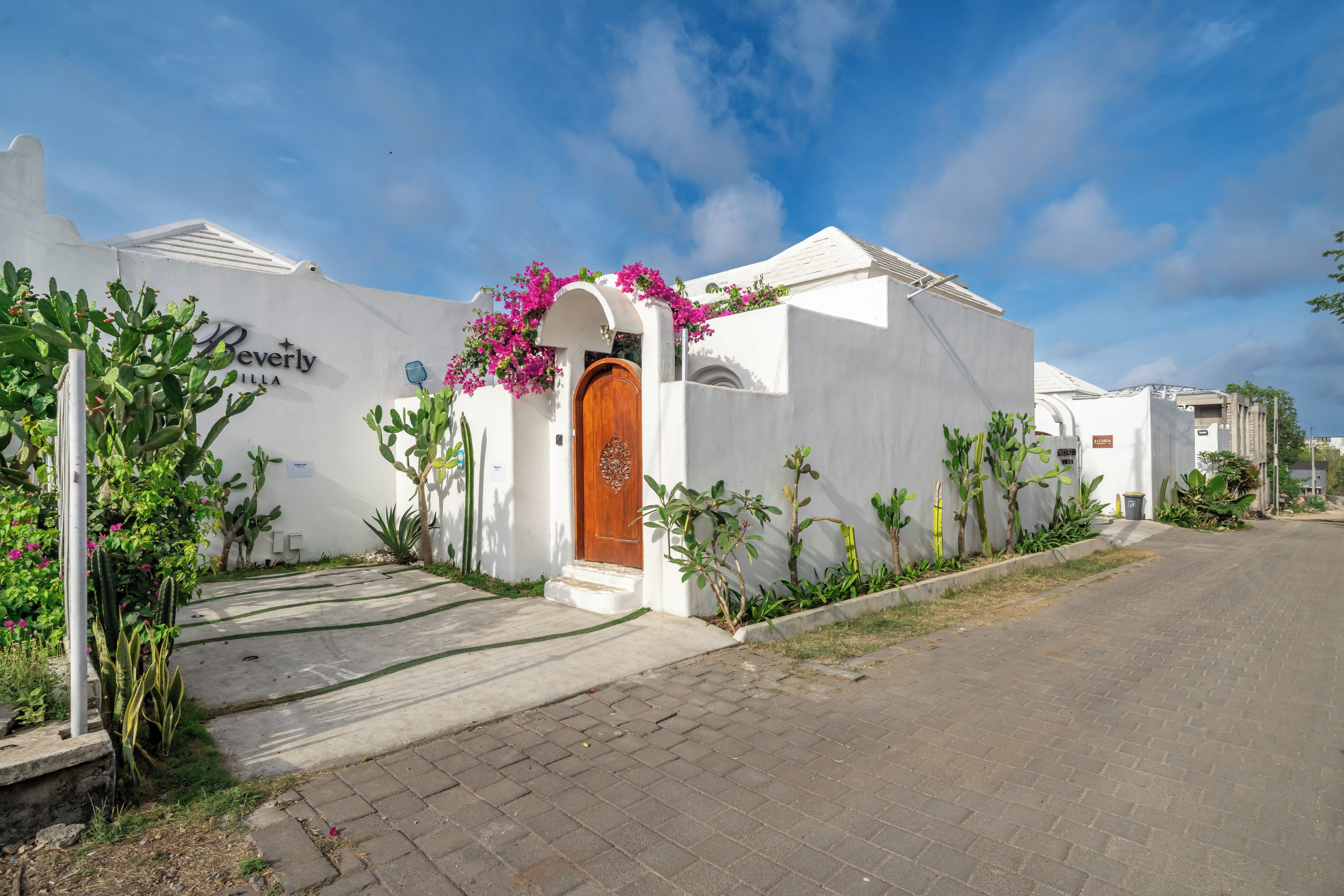 Un edificio blanco con puerta de madera y flores rosadas, rodeado de cactus y plantas.
