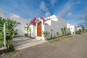 Un edificio blanco con puerta de madera y flores rosadas, rodeado de cactus y plantas.