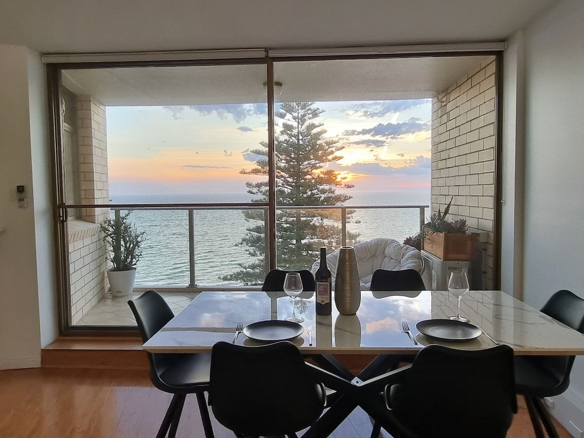 Uma área de refeições com uma mesa posta para dois, com vista para o mar e árvores ao pôr do sol.