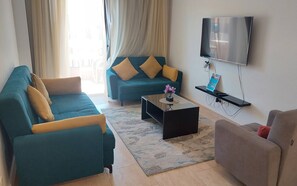 A living room with a teal sofa, a coffee table, a TV mounted on the wall, and a small shelf with a remote control. - Luxurious Chalet in Red Sea Governorate with Stunning Sea Views (Red Sea Governorate)