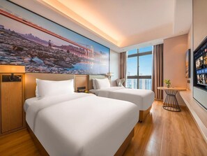 Et hotelværelse med to senge, et stort vindue, et bybillede som maleri og et fjernsyn på væggen.