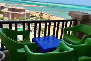 Uma varanda com cadeiras de plástico verde e mesa azul, com vista para uma praia, edifícios e um mar azul cristalino.