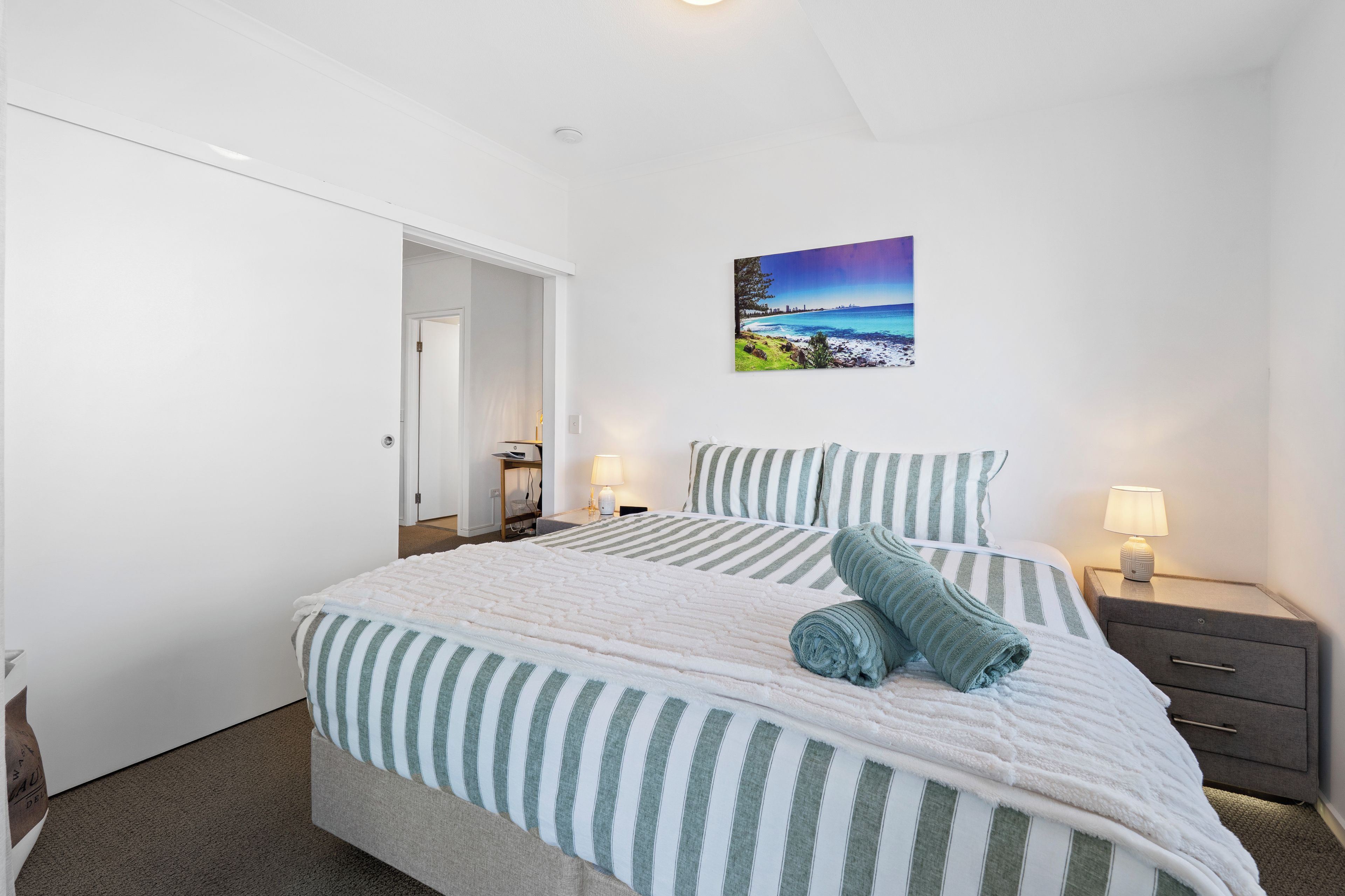 A bedroom with a bed, pillows, a nightstand, a lamp, and a painting on the wall.