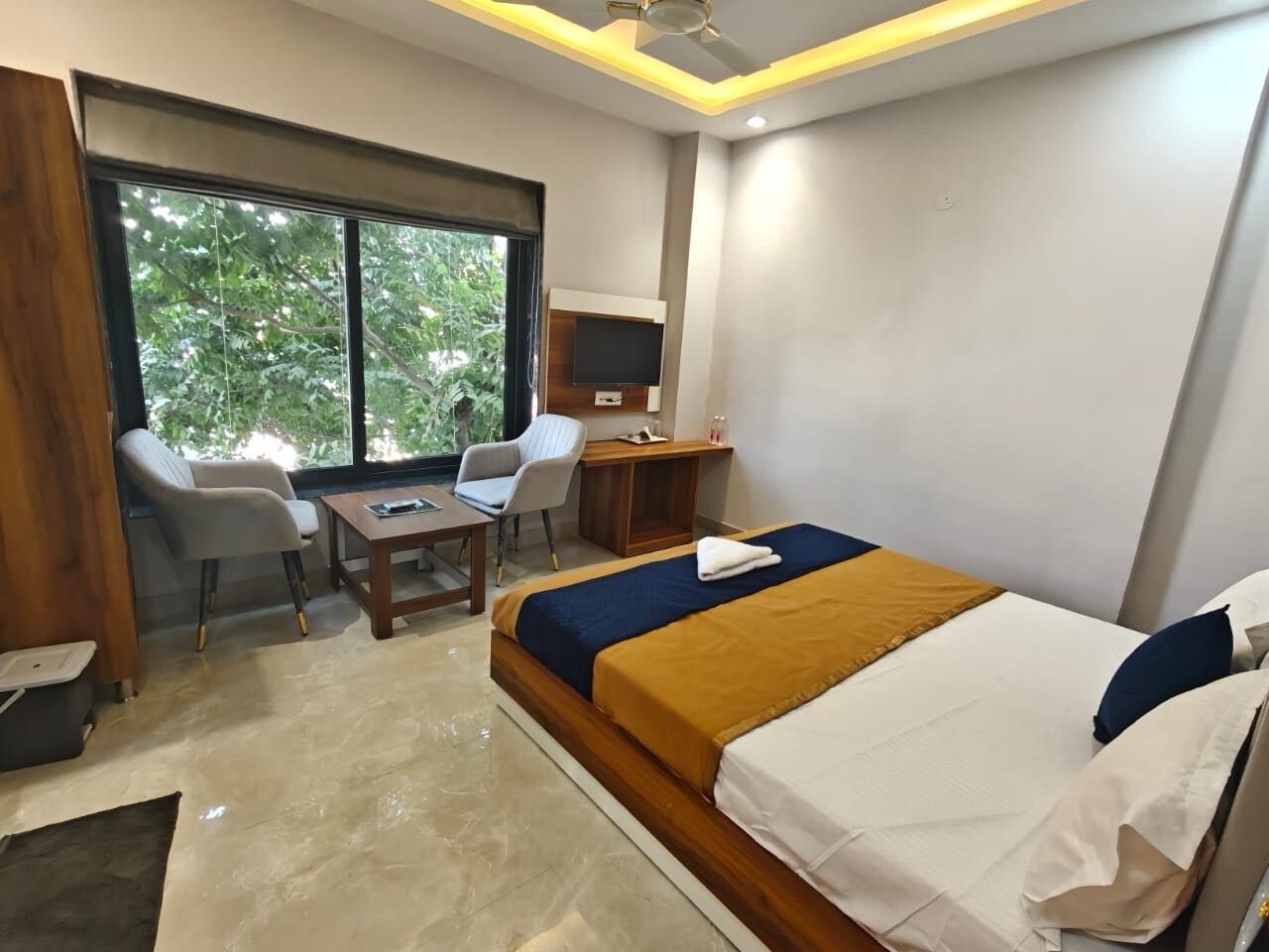 A hotel room with a bed, a desk, two chairs, a television, and a window with a view of greenery.