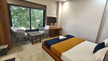 A hotel room with a bed, a desk, two chairs, a television, and a window with a view of greenery.