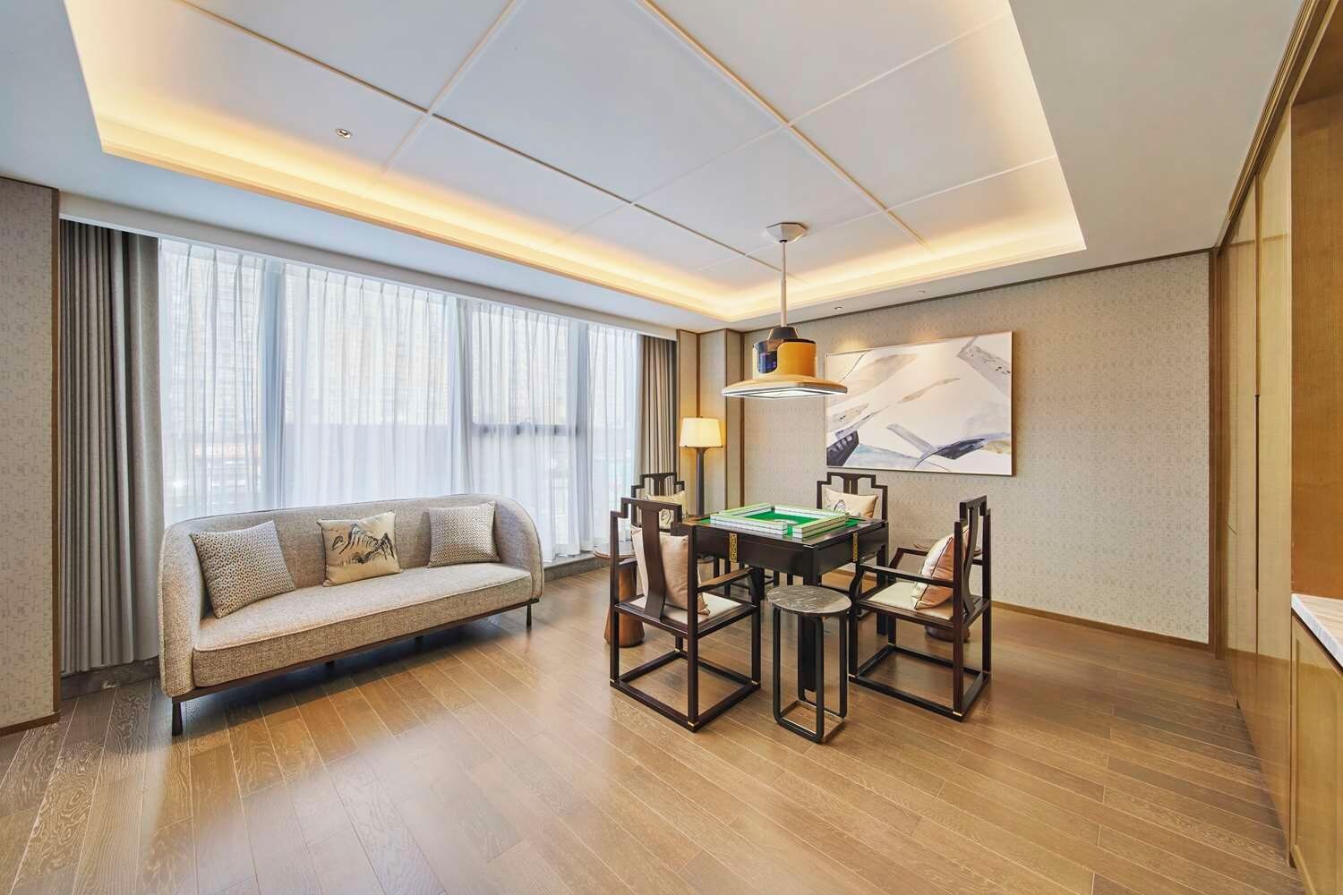 A modern hotel room with a sofa, dining table, and chairs, a large window with curtains, and a ceiling light fixture.