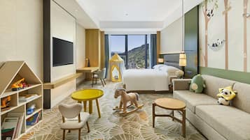 Habitación de hotel con cama, sofá, escritorio, silla, una mesita pequeña, estantería y un ventanal con vistas a las montañas.