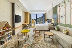 Habitación de hotel con cama, sofá, escritorio, silla, una mesita pequeña, estantería y un ventanal con vistas a las montañas.