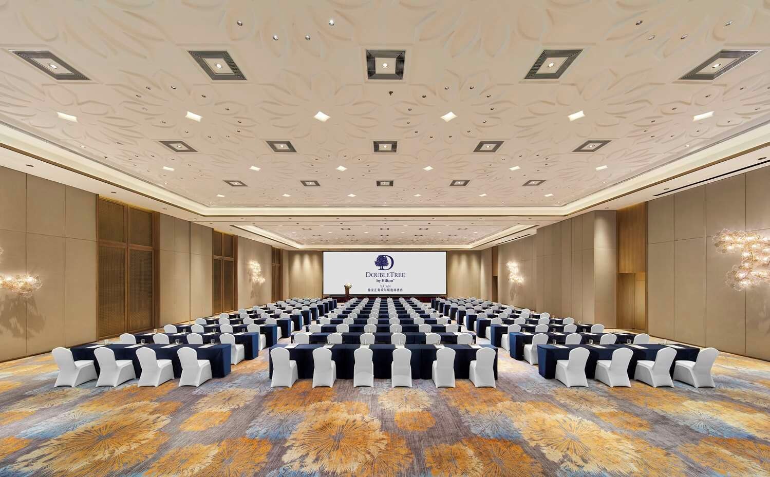 A conference room with rows of tables and chairs, a large screen at the front, and a patterned carpet.