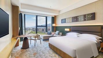 A hotel room with a large bed, a desk with a chair, a sofa, and a large window offering a city view.