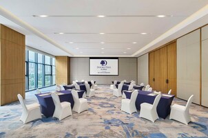 Un salón de conferencias con una pantalla grande que muestra el logotipo de DoubleTree by Hilton, sillas blancas y piso alfombrado.