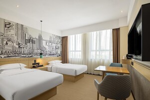 A hotel room with two beds, a desk, a chair, a TV, and a cityscape mural on the wall.