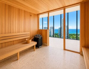 A sauna room with wooden benches, a trash bin, and a large window offering a view of buildings and the ocean. - Luxury 2BR | Ocean Views | Level 50 Broadbeach (Broadbeach)