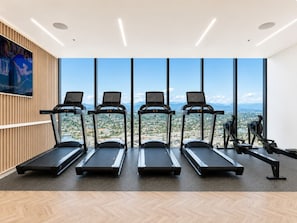 A gym with treadmills and rowing machines, large windows with a city view, and a wooden wall with a poster. - Luxury 2BR | Ocean Views | Level 50 Broadbeach (Broadbeach)