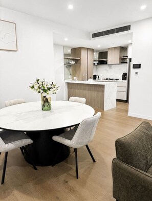A modern kitchen with a central island, white walls, and a dining area with a round table and chairs. - Luxury 2BR | Ocean Views | Level 50 Broadbeach (Broadbeach)