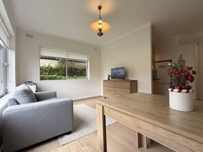 A modern living room with a sofa, a wooden dining table, a TV on a cabinet, and a window with a view of greenery. - Cremorne Point Harbour Escape (Cremorne Point)